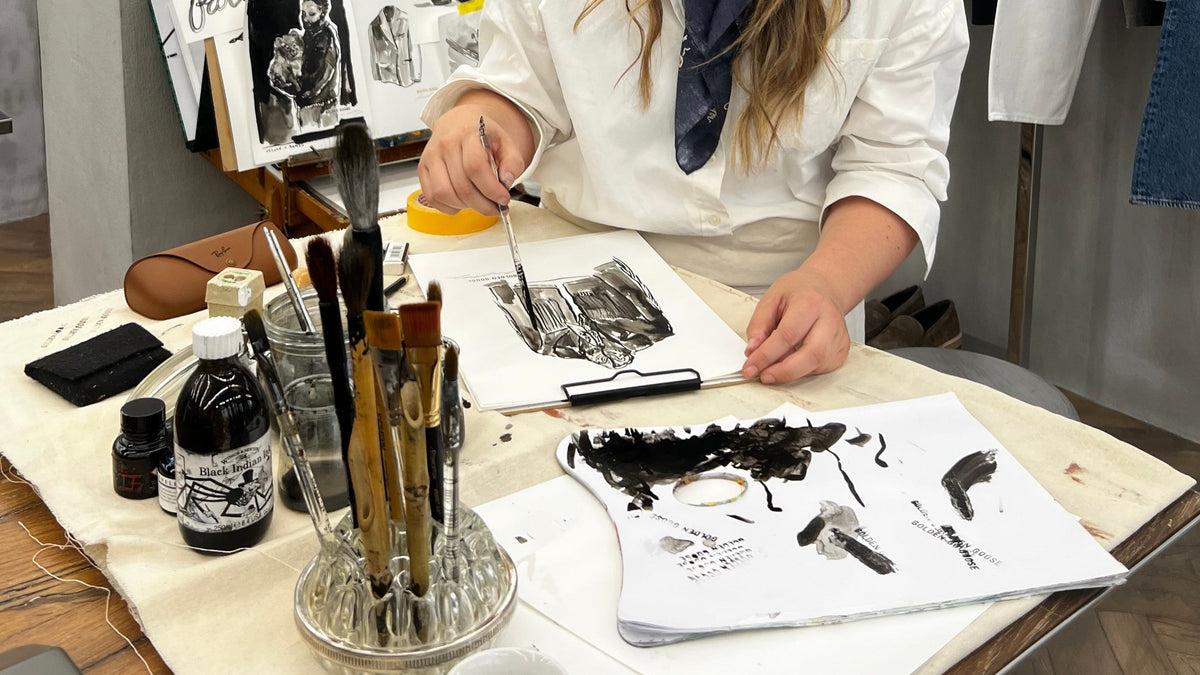 A woman with long brown hair sits at a wooden table in a creative studio, smiling as she works on fashion sketches. She is wearing a white long-sleeve shirt and a navy blue patterned scarf.